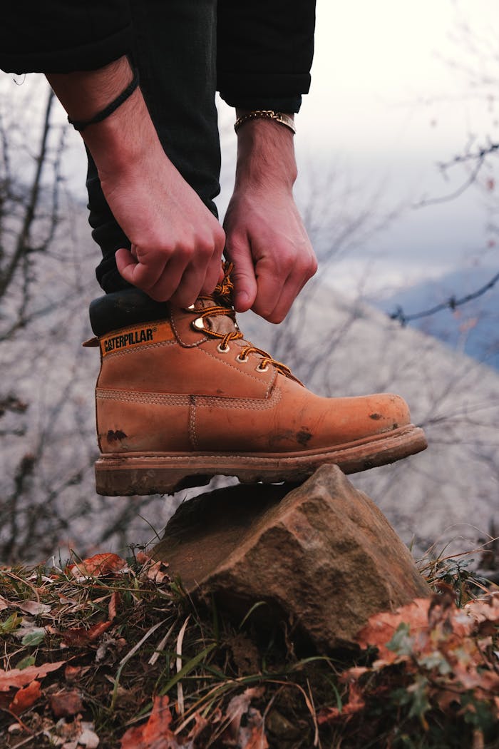 Person tying brown hiking boot on a rock outdoors, focusing on footwear.