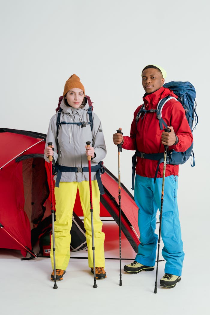 Two hikers with trekking poles and backpacks stand near a red tent, ready for adventure.